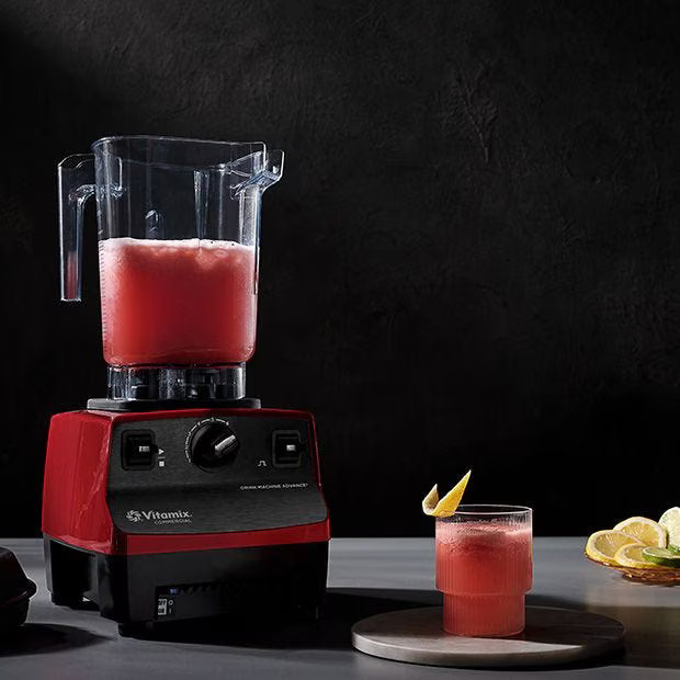 Un blender Vitamix roșu umplut cu un smoothie roz se află pe un tejghea, lângă un pahar cu aceeași băutură, cu o garnitură de lămâie. Lămâile feliate sunt vizibile pe o farfurie în dreapta. Fundalul este întunecat și minimalist.