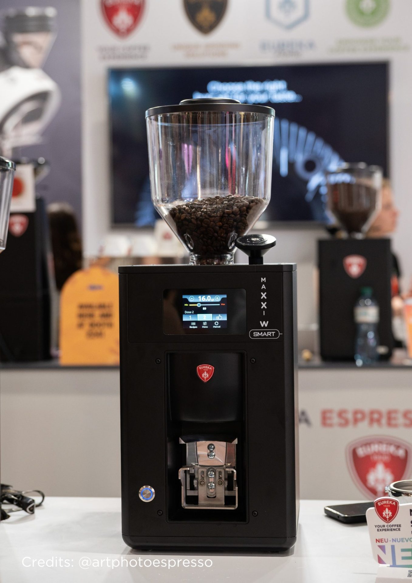 Un espressor modern plin cu boabe de cafea este expus pe un tejghea la o expoziție. Pe fundal se văd bannere neclare și oameni. Credit: @artphotoespresso.
