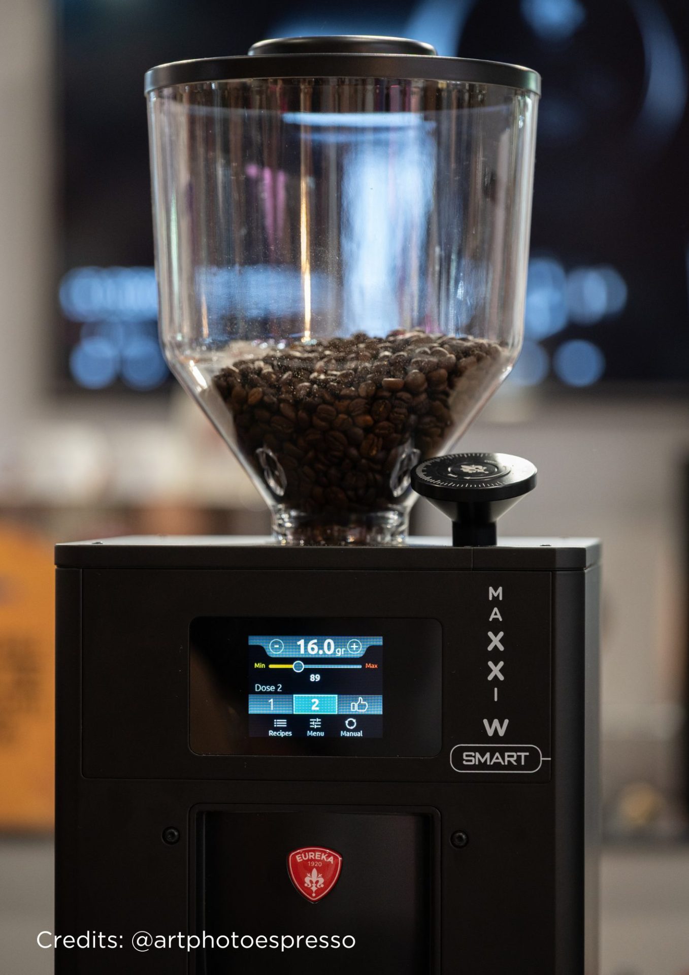 Un prim plan al unui râșniță de cafea neagră plină cu boabe de cafea. Afișajul digital de pe partea frontală arată setările de măcinare și cuvântul SMART. Mașina de măcinat este marcată cu un logo Eureka roșu. Credits: @artphotoespresso.