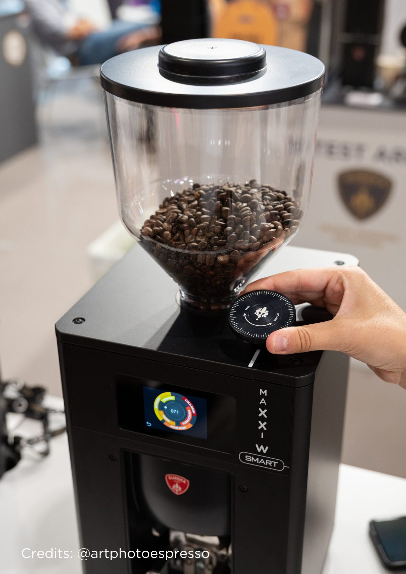 O mână reglează setarea de măcinare la un măcinător de cafea modern, plin cu boabe de cafea. Moara are un afișaj digital și un rezervor mare și transparent deasupra.