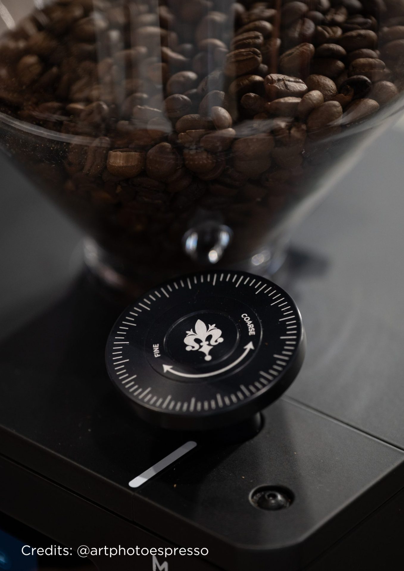 Prim plan al unei râșnițe de cafea plină cu boabe de cafea. Accentul se pune pe cadranul de măcinare, etichetat Fine și Course cu săgeți și un simbol al florii de crin. Credit foto: @artphotoespresso.