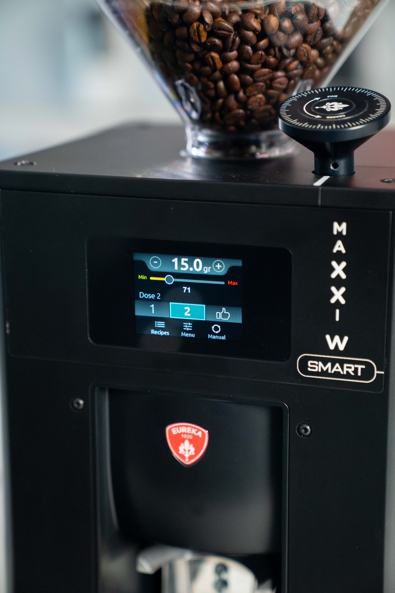 Prim plan al unui măcinător de cafea Eureka negru cu un afișaj digital care arată setările pentru cantitatea de măcinare, doză și moduri. Recipientul superior este umplut cu boabe de cafea, iar cadranele de reglare sunt vizibile.