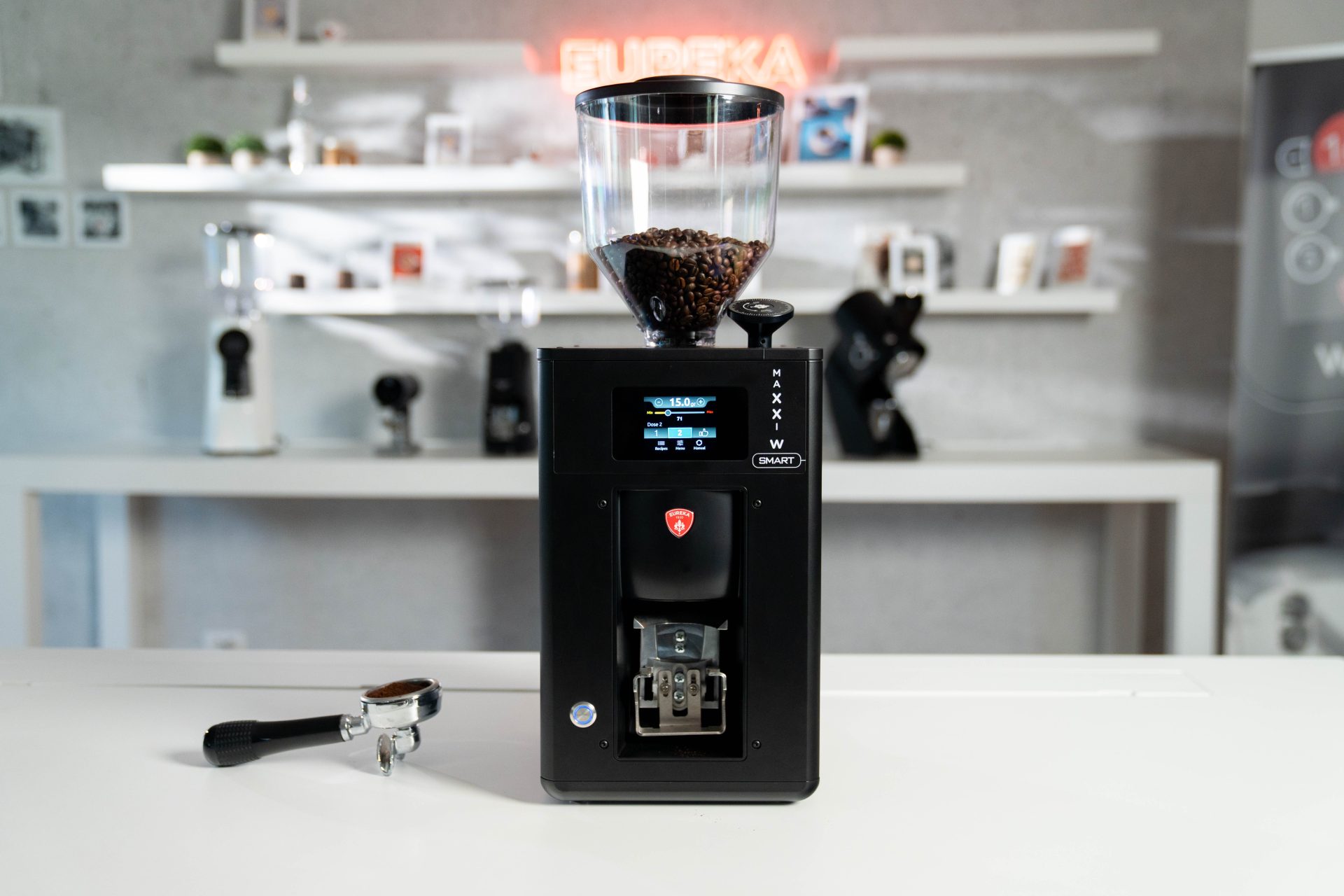O râșniță de cafea neagră plină cu boabe de cafea se află pe un blat alb. Un portafilter este așezat în apropiere, iar rafturile cu echipamente de cafea sunt vizibile în fundal.